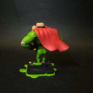 K&ouml;nnte beinhalten: Figur eines Charakters in gr&uuml;nem Outfit und rotem Umhang, stehend auf einer schwarzen Basis mit gr&uuml;nem Schleim. Die Figur hat einen beigen Hut und ist mit den Armen an den Seiten posiert. Der Hintergrund ist dunkel.