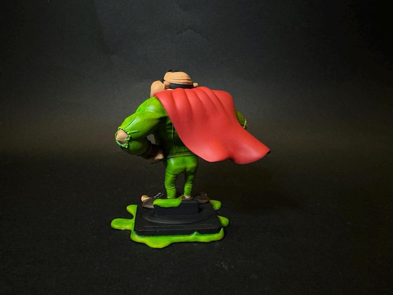 K&ouml;nnte beinhalten: Figur eines Charakters in gr&uuml;nem Outfit und rotem Umhang, stehend auf einer schwarzen Basis mit gr&uuml;nem Schleim. Die Figur hat einen beigen Hut und ist mit den Armen an den Seiten posiert. Der Hintergrund ist dunkel.