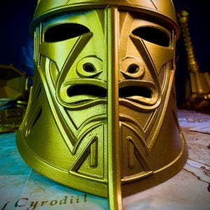 K&ouml;nnte beinhalten: Ein goldfarbener Helm mit geteiltem Gesicht und komplizierten geometrischen Mustern. Der Helm hat Augen- und Mund&ouml;ffnungen sowie eine vertikale Teilung in der Mitte. Im Hintergrund sind eine Karte und ein dunkelblauer Hintergrund zu sehen.