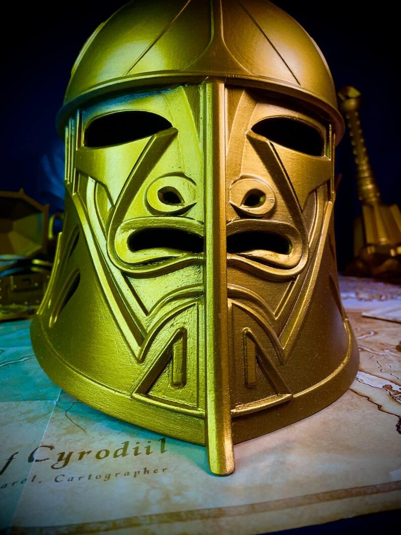 K&ouml;nnte beinhalten: Ein goldfarbener Helm mit geteiltem Gesicht und komplizierten geometrischen Mustern. Der Helm hat Augen- und Mund&ouml;ffnungen sowie eine vertikale Teilung in der Mitte. Im Hintergrund sind eine Karte und ein dunkelblauer Hintergrund zu sehen.