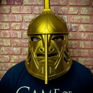 K&ouml;nnte beinhalten: Ein goldfarbener Helm mit einem markanten Gesichtsdesign und einem vertikalen Kamm. Der Helm wird &uuml;ber einem dunkelblauen T-Shirt mit dem Aufdruck "GAME OF THRONES" getragen. Der Hintergrund ist eine Backsteinmauer.