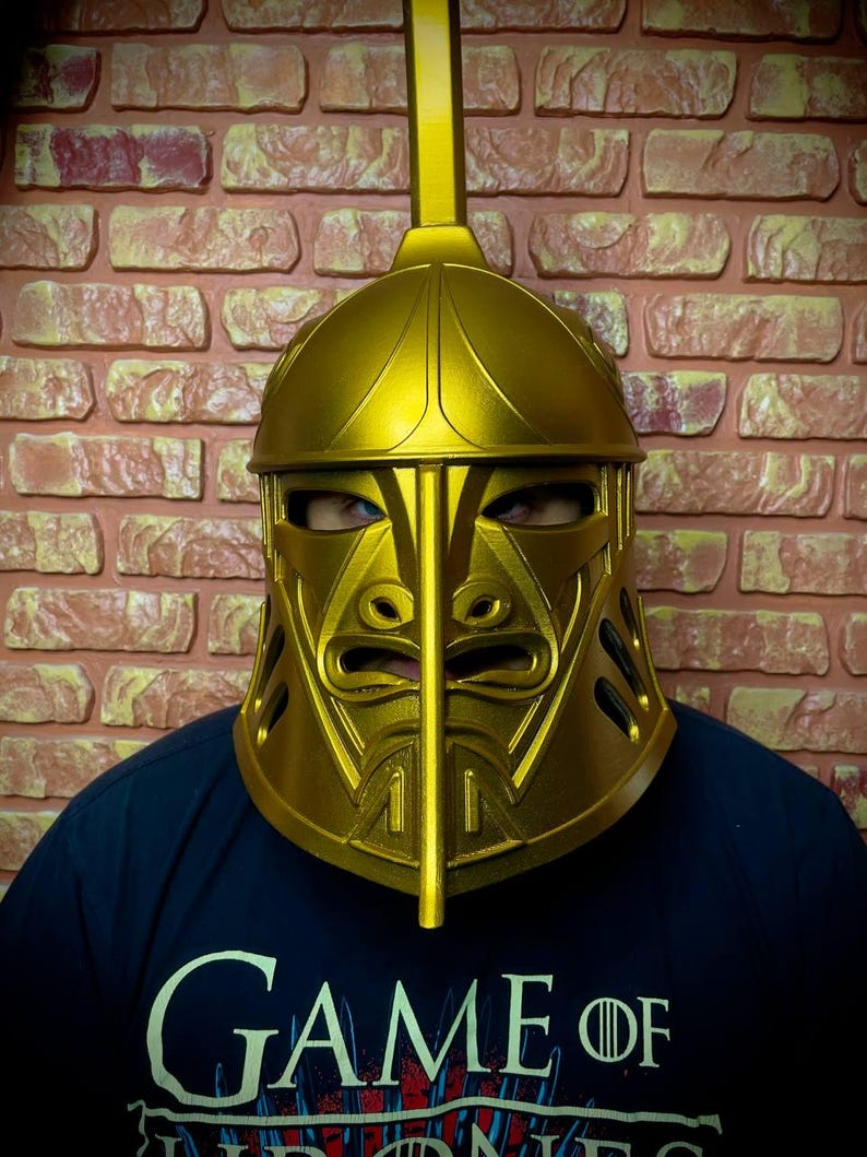 K&ouml;nnte beinhalten: Ein goldfarbener Helm mit einem markanten Gesichtsdesign und einem vertikalen Kamm. Der Helm wird &uuml;ber einem dunkelblauen T-Shirt mit dem Aufdruck "GAME OF THRONES" getragen. Der Hintergrund ist eine Backsteinmauer.