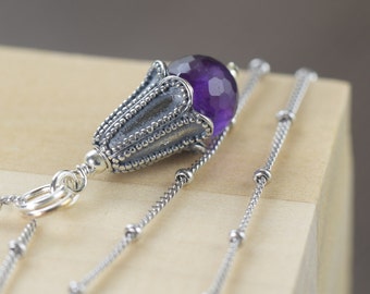 May include: A silver necklace with a detailed pendant featuring a purple amethyst gemstone. The pendant is shaped like a flower bud.