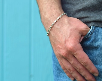 May include: A silver chain bracelet with a box chain design. The bracelet is on a person's wrist.