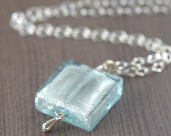 May include: A silver chain necklace with a square pendant made of light blue glass with a silver finish. The pendant has a small silver drop hanging from it.