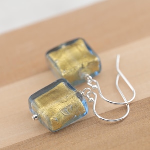 May include: A pair of square drop earrings with a gold leaf design encased in clear blue glass. The earrings have silver-colored ear wires. The earrings are displayed on a light brown wooden surface.