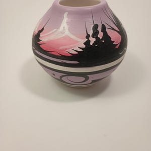 May include: A small, round, hand-painted ceramic vase. The vase features a lavender background with a painted landscape scene in shades of pink, white, and black. The design includes a black silhouette of trees and a white and black geometric pattern.