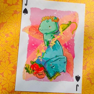 May include: A Jack of Spades playing card with a watercolour illustration of a teal creature wearing a flower crown. The artwork is set against a pink and orange backdrop. The card's suit symbol and "J" are in the corners.