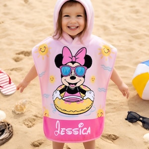 May include: A pink hooded beach towel featuring Minnie Mouse wearing sunglasses and a floatie. The towel has the name "Jessica" printed on it. A beach ball, sunglasses, and flip-flops are on the sand.