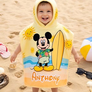 Puede incluir: Una toalla de playa con capucha amarilla con Mickey Mouse con una tabla de surf, soles y olas del oc&eacute;ano. La toalla tiene el nombre "Anthony" impreso. Otros accesorios de playa incluyen una pelota de playa, gafas de sol y chanclas.