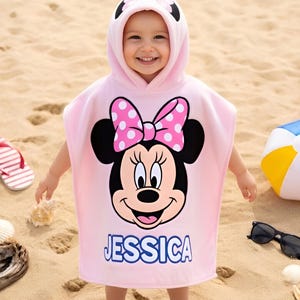 May include: A child wearing a pink hooded towel featuring Minnie Mouse with a polka dot bow and the name "JESSICA". The towel is on a sandy beach, with flip-flops, a beach ball, and sunglasses nearby.