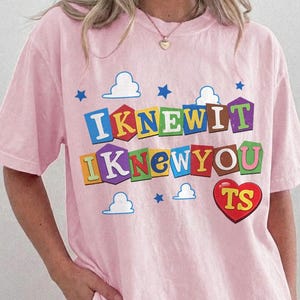 May include: A light pink short-sleeve t-shirt featuring a graphic print with the text "IKNEWIT IKNEWYOU" in colorful block letters resembling toy blocks. The design includes small blue stars, white clouds, and a red heart with the letters "TS".
