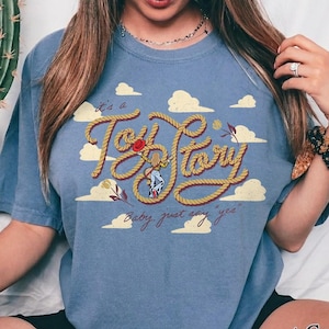May include: A blue short-sleeve t-shirt featuring a graphic print of a rope shaped into the text "Toy Story" with a small cowboy figure in the center. The design includes yellow clouds and the cursive text "it's a" and "Baby, just say "yes"" in brown.