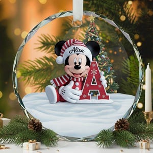 Puede incluir: Una bola de Navidad redonda y transparente con Mickey Mouse con gorro y bufanda de Papá Noel, sosteniendo la letra roja "A". El nombre "Alisa" está en el sombrero. La bola cuelga frente a un árbol de Navidad y velas.