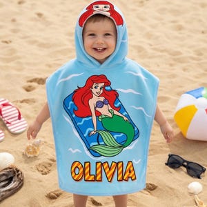 K&ouml;nnte beinhalten: Ein hellblaues Strandtuch mit Kapuze, das ein Cartoon-Meerjungfrau-Design und den Namen "OLIVIA" in Gelb zeigt. Das Handtuch hat eine Kapuze mit einem Meerjungfrau-Design. Das Bild zeigt auch einen Strandball, eine Sonnenbrille und Flip-Flops.