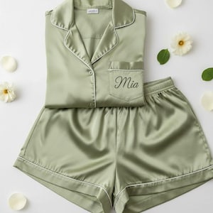Puede incluir: Conjunto de pijama de satén verde salvia con ribete blanco y el nombre "Mia" bordado en el bolsillo. El conjunto incluye una camisa con botones y pantalones cortos a juego. El conjunto está rodeado de pétalos de flores blancas y hojas verdes.