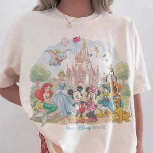 May include: An ivory short-sleeve t-shirt featuring a colorful graphic print of various Disney characters in front of a castle. The text "Magic Kingdom" and "Walt Disney World" is printed below the characters. The shirt has a crew neckline.