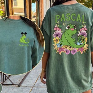 Peut inclure: Un t-shirt vert clair avec au dos un motif de caméléon vert entouré de fleurs colorées et le texte PASCAL. La poitrine affiche un plus petit caméléon vert portant des oreilles de souris avec le texte Name inscrit juste en dessous.
