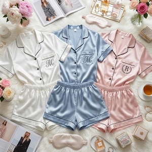 May include: Three sets of satin pajamas in white, light blue, and pink, each with a matching shorts set. The pajamas have black trim and embroidered initials. The image includes a tray of perfumes, flowers, and a sleep mask.