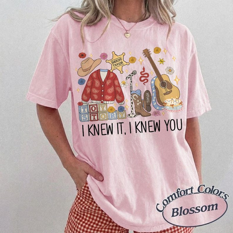 May include: A light pink t-shirt featuring a graphic print with a cowboy hat, a red paisley cardigan, a sheriff badge, a microphone, a brown acoustic guitar, and alphabet blocks spelling TOY STORY. The text reads I KNEW IT, I KNEW YOU.