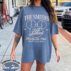 May include: Blue jean comfort color t-shirt with the text "THE SMITHS" and "Just Married" in white. The shirt also features a Mickey Mouse bow design and the words "Newlyweds Club" and "MAGIC KING DOM".