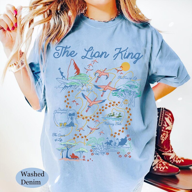 May include: A light blue t-shirt with a graphic print of a map of the Lion King movie. The map includes locations like Pride Rock, The Watering Hole, and The Circle of Life. The text "The Lion King" is printed at the top of the map.