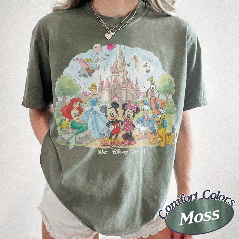 May include: A moss green t-shirt featuring a graphic print of various Disney characters including Mickey Mouse, Minnie Mouse, Donald Duck, Goofy, Pluto, Cinderella, Ariel, and Dumbo in front of a castle with the text Magic Kingdom Walt Disney World.