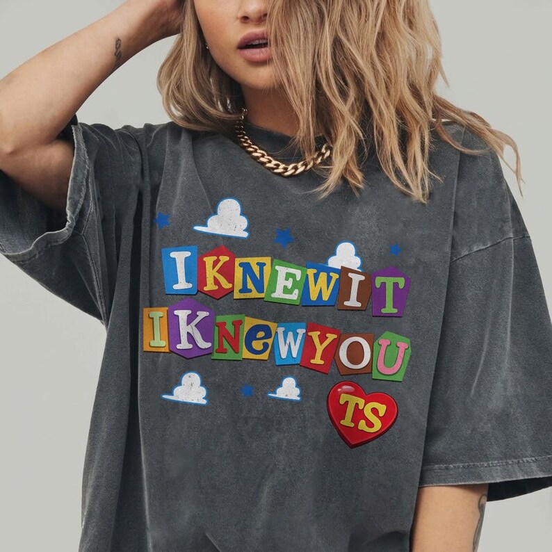 May include: A charcoal gray short-sleeve t-shirt featuring a graphic print with the text I KNEW IT I KNEW YOU in colorful, block-letter cutouts resembling ransom notes. The design includes white cloud shapes, small blue stars, and a red heart with TS.