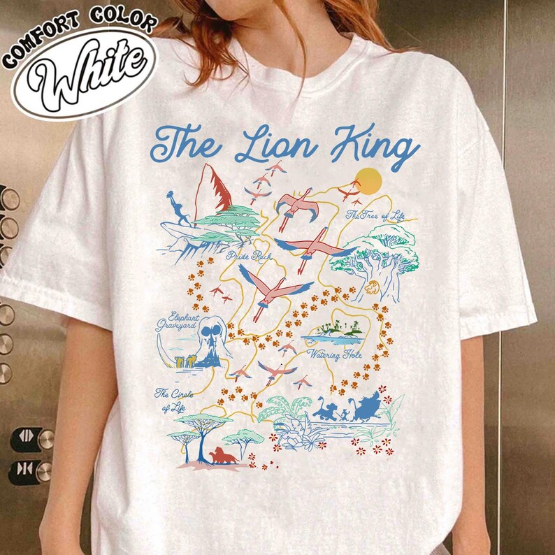 May include: White t-shirt with a colorful map of the Lion King's Pride Lands. The map includes locations like Pride Rock, the Watering Hole, and the Elephant Graveyard. The text "The Lion King" is printed in blue at the top of the map.