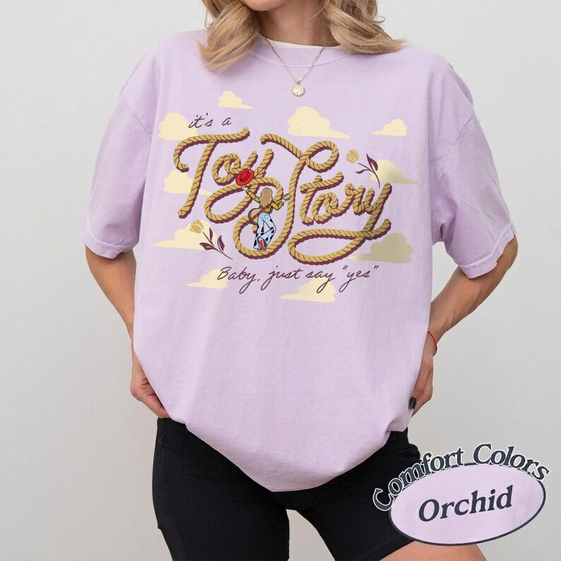 May include: A light purple t-shirt featuring a graphic of a rope shaped into the text Toy Story, with a small illustration of a cowgirl character in the center. The text it's a and Baby, just say yes is printed in a cursive font with clouds.
