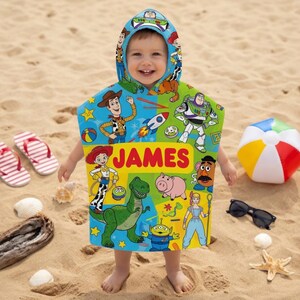 Puede incluir: Una toalla de playa con capucha para ni&ntilde;os con personajes de Toy Story y el nombre "JAMES". La toalla es colorida y presenta im&aacute;genes de Woody, Buzz Lightyear y otros personajes. Un bal&oacute;n de playa, gafas de sol y chanclas est&aacute;n en la arena.