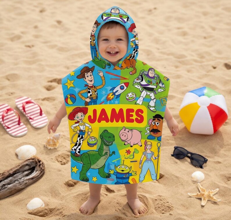 Puede incluir: Una toalla de playa con capucha para ni&ntilde;os con personajes de Toy Story y el nombre "JAMES". La toalla es colorida y presenta im&aacute;genes de Woody, Buzz Lightyear y otros personajes. Un bal&oacute;n de playa, gafas de sol y chanclas est&aacute;n en la arena.