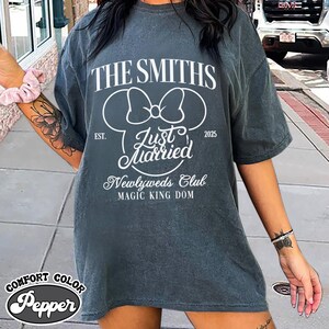 May include: A gray Comfort Colors t-shirt with the text "THE SMITHS" and "Just Married" in white. The design includes a Mickey Mouse head silhouette and the words "Newlyweds Club" and "MAGIC KING DOM". The year "2025" is also printed on the shirt.