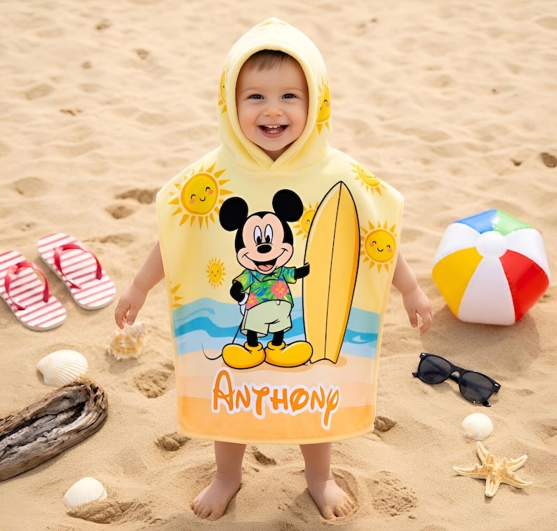 Puede incluir: Una toalla de playa con capucha amarilla con Mickey Mouse con una tabla de surf, soles y olas del oc&eacute;ano. La toalla tiene el nombre "Anthony" impreso. Otros accesorios de playa incluyen una pelota de playa, gafas de sol y chanclas.