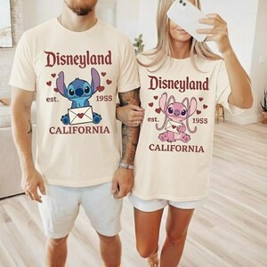 Op de afbeelding: Twee cr&egrave;mekleurige T-shirts met het Disneyland-logo en een stripfiguur met een envelop. De shirts hebben ook de tekst "est. 1955 CALIFORNIA". E&eacute;n shirt heeft een blauw karakter, de andere een roze karakter.