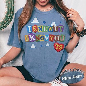May include: A blue short-sleeve t-shirt featuring a graphic print with the text I KNEW IT I KNEW YOU TS in colorful block letters. The design includes white cloud shapes, small blue stars, and a red heart shape containing the letters TS.
