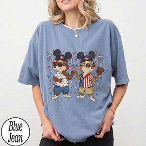 May include: A light blue t-shirt featuring a cartoon illustration of two chipmunks wearing sunglasses and Mickey Mouse ears, holding American flags and Mickey Mouse-shaped treats. The shirt has a round neckline and short sleeves. The text "Blue Jean" is in a circle at the bottom.