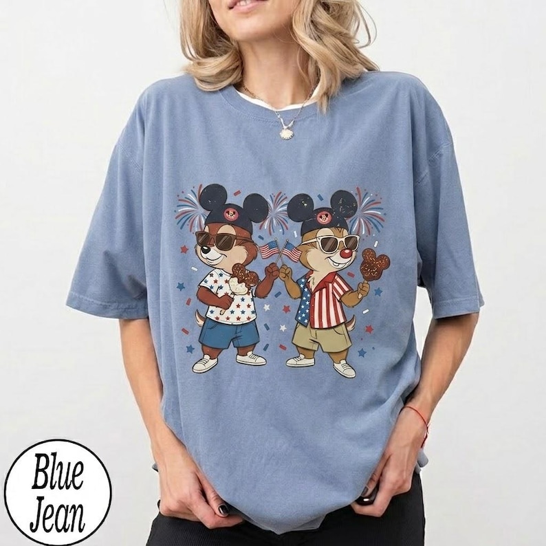 May include: A light blue t-shirt featuring a cartoon illustration of two chipmunks wearing sunglasses and Mickey Mouse ears, holding American flags and Mickey Mouse-shaped treats. The shirt has a round neckline and short sleeves. The text "Blue Jean" is in a circle at the bottom.
