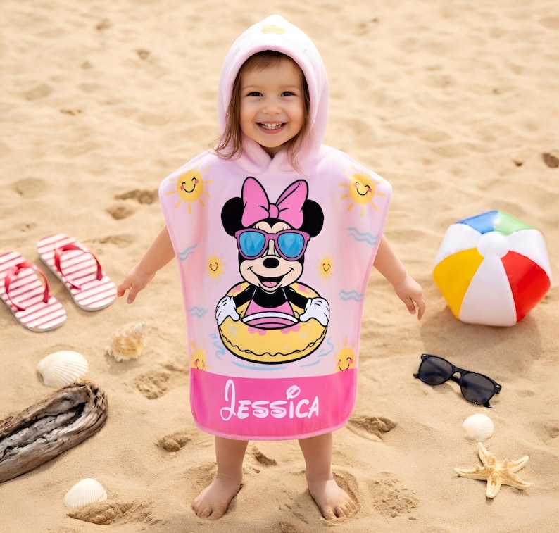May include: A pink hooded beach towel featuring Minnie Mouse wearing sunglasses and a floatie. The towel has the name "Jessica" printed on it. A beach ball, sunglasses, and flip-flops are on the sand.