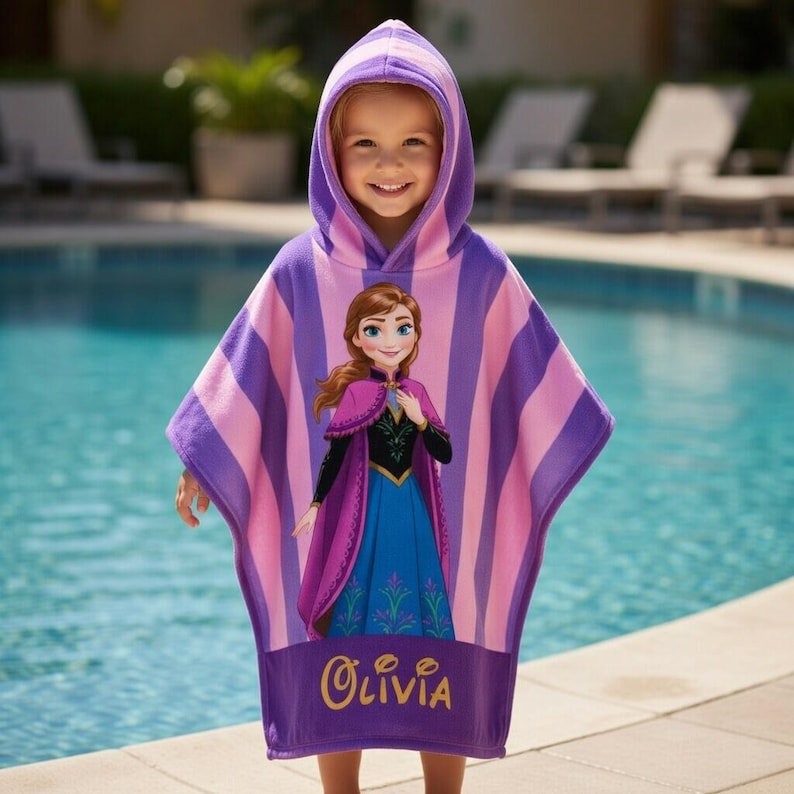 Puede incluir: Una toalla de playa con capucha para ni&ntilde;os con una imagen de dibujos animados de Anna de Frozen. La toalla tiene rayas rosas y moradas. El nombre "Olivia" est&aacute; bordado en dorado en la parte inferior.