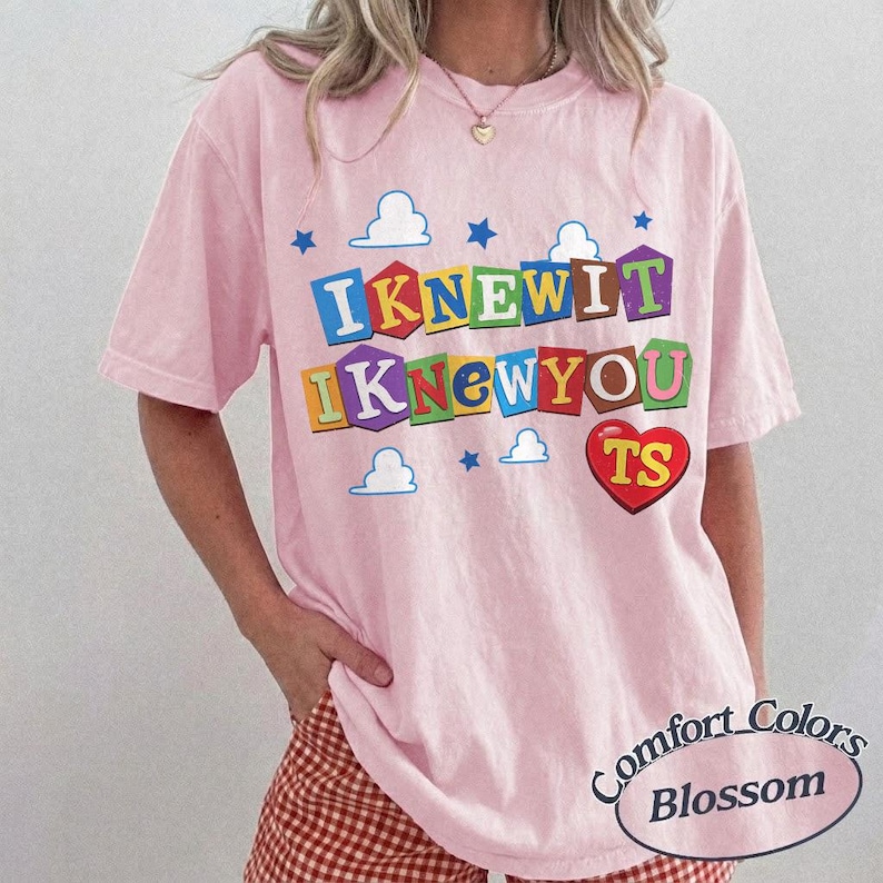 May include: A light pink short-sleeve t-shirt featuring a graphic print with the text "IKNEWIT IKNEWYOU" in colorful block letters resembling toy blocks. The design includes small blue stars, white clouds, and a red heart with the letters "TS".