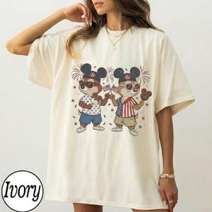 May include: Ivory-colored t-shirt featuring a cartoon graphic of two chipmunks wearing sunglasses and Mickey Mouse ears, holding American flags and ice cream. The chipmunks are dressed in patriotic outfits, with fireworks and star accents.