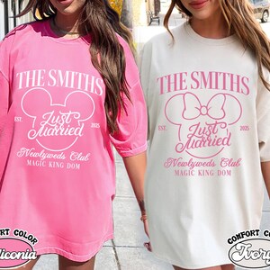 May include: Two oversized t-shirts, one pink and one ivory, featuring a Disney-themed design. The design includes the text "The Smiths," "Just Married," and "Newlyweds Club," with a Mickey Mouse head or bow. The shirts are labeled "Comfort Color."