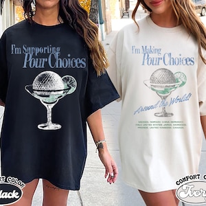 Könnte beinhalten: Schwarze und elfenbeinfarbene Comfort Color T-Shirts mit einem Cocktail-Motiv. Das Motiv zeigt einen Golfball in einem Martiniglas mit dem Text "I'm Supporting Pour Choices" und "I'm Making Pour Choices Around the World!"
