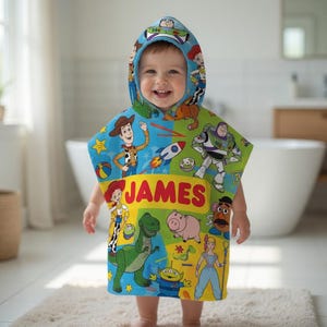 Puede incluir: Una toalla con capucha y colorida con personajes de Toy Story. La toalla es azul, verde y amarilla, con el nombre "JAMES" impreso en letras rojas grandes.