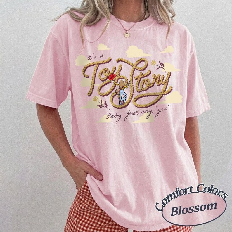 May include: A light pink short-sleeve t-shirt featuring a graphic of a rope shaped into the text Toy Story, with a small illustration of a cowgirl character, yellow clouds, and the text Baby, just say yes in a cursive script font.