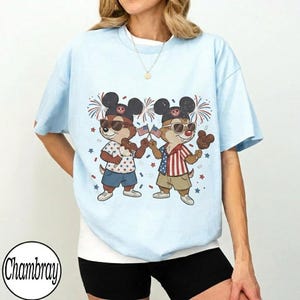 May include: Light blue t-shirt featuring a cartoon illustration of two chipmunks wearing sunglasses and Mickey Mouse ears. The chipmunks are holding American flags and Mickey Mouse-shaped ice cream treats, with fireworks and stars in the background.