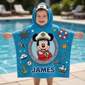 Op de afbeelding: Een blauwe badponcho met een Mickey Mouse-ontwerp. De poncho heeft nautische afbeeldingen, waaronder een scheepsstuur, anker en reddingsboei. De naam "JAMES" is in witte blokletters op de poncho gedrukt.