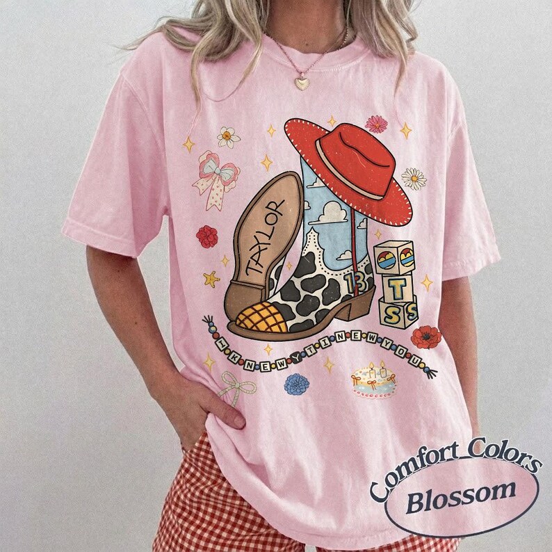 May include: A light pink short-sleeve t-shirt featuring a graphic print of a cowboy boot with a cow print pattern, a red hat, the name Taylor, the number 13, building blocks, a beaded necklace, and small flowers on the front center.