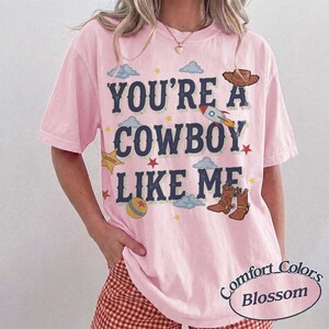 May include: A light pink short-sleeve t-shirt featuring the dark blue text "YOU'RE A COWBOY LIKE ME" surrounded by illustrations of a brown cowboy hat, a rocket ship, a yellow sheriff star, a colorful ball, and brown cowboy boots with clouds.